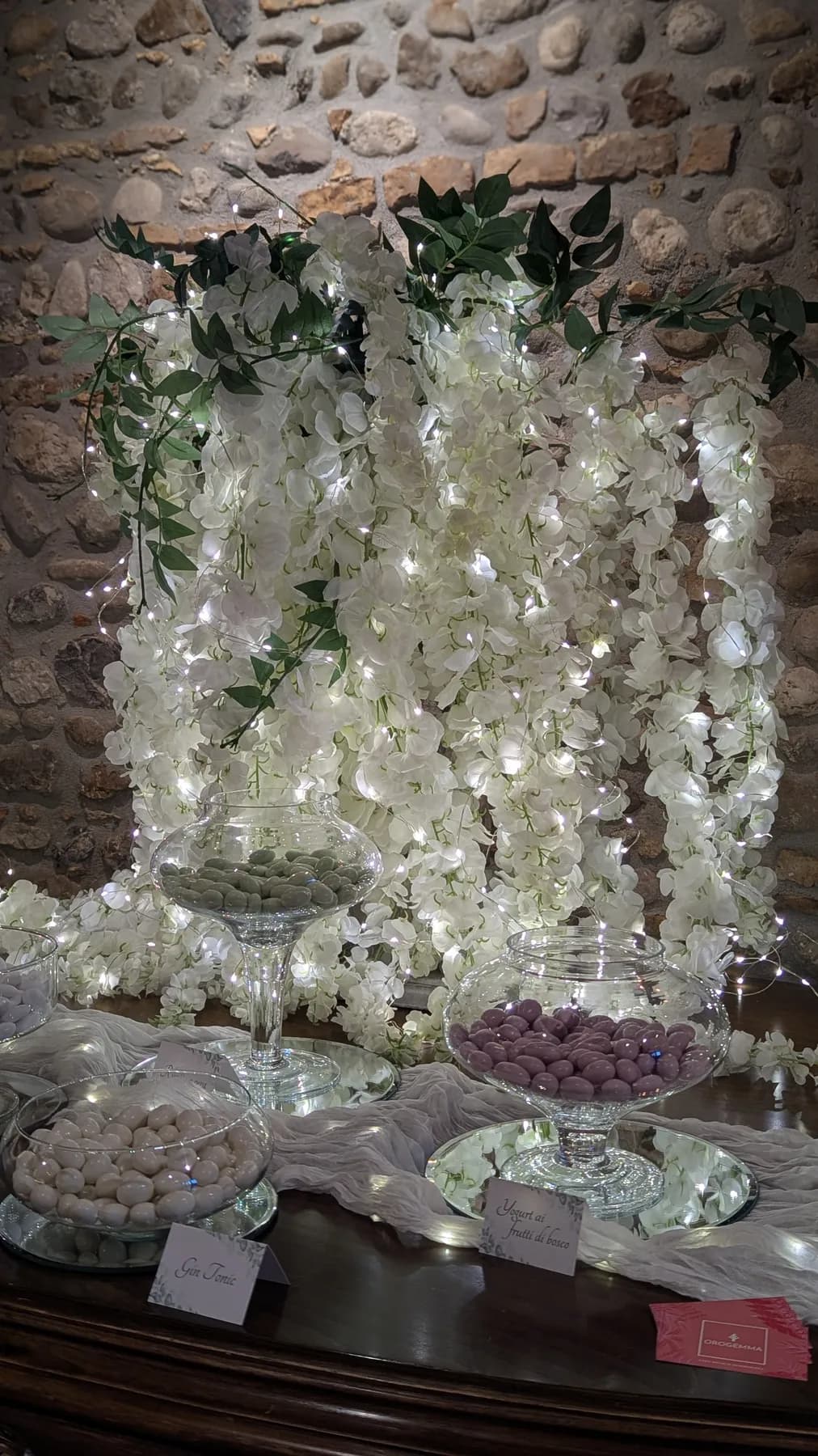Allestimento con cascate di fiori bianchi illuminate e vasi di confetti